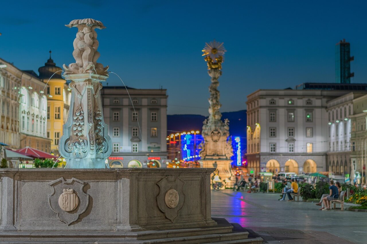 Linz, Rakousko: Náměstí s fontánou a sloupem Nejsvětější Trojice v noci. Turistická destinace, architektura.