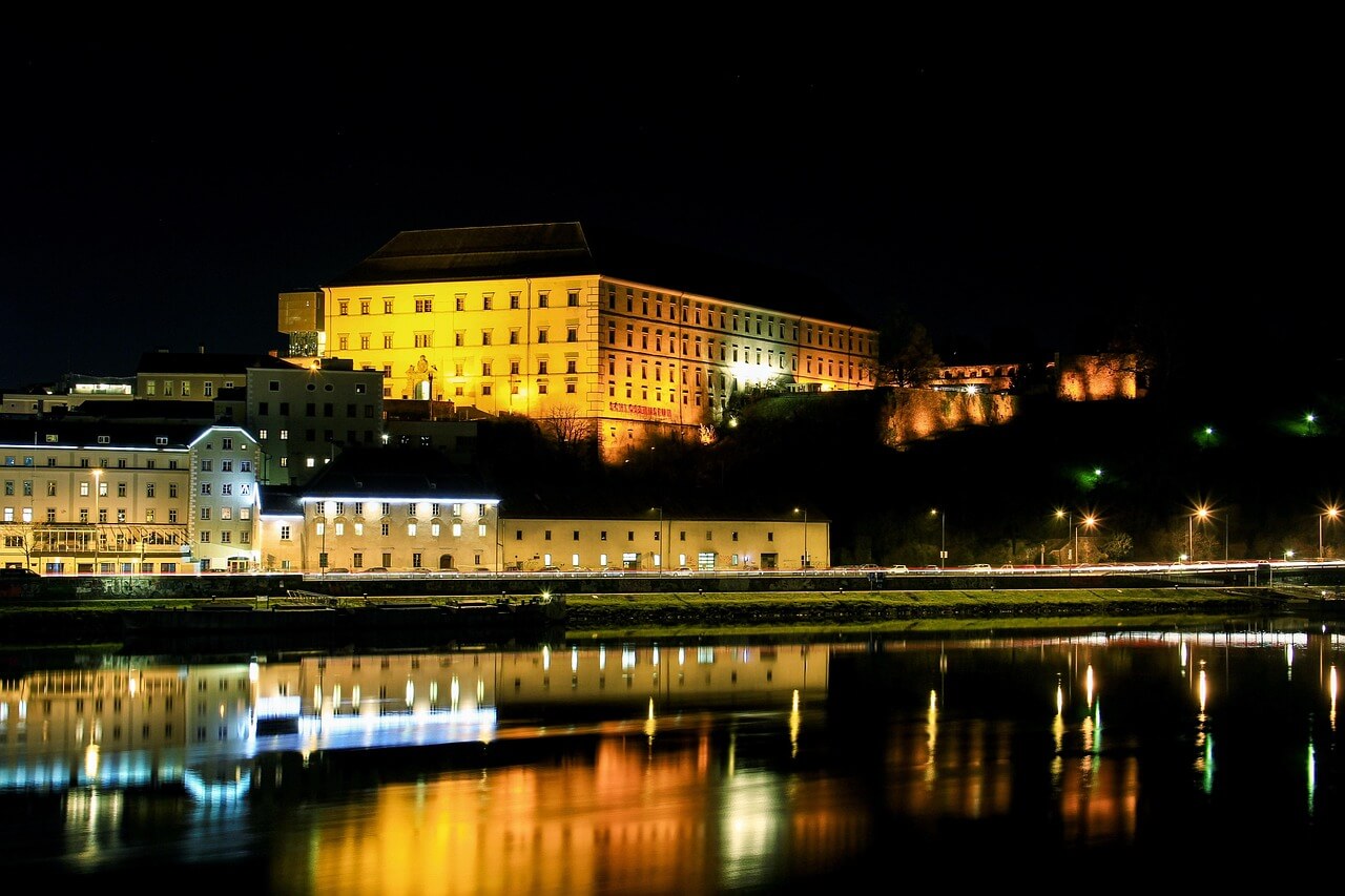 Noční pohled na hrad, budovy a řeku. Světla se odrážejí ve vodě. Architektura, cestování, linz
