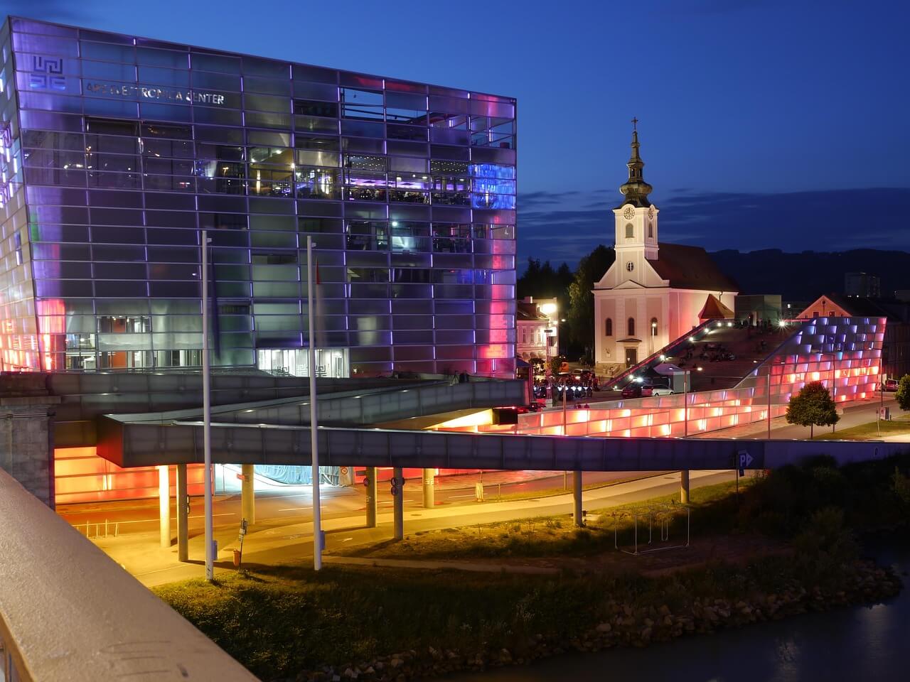 Architektonické budovy v Linci v noci. Moderní centrum a kostel s věží. Městská krajina, noční osvětlení.