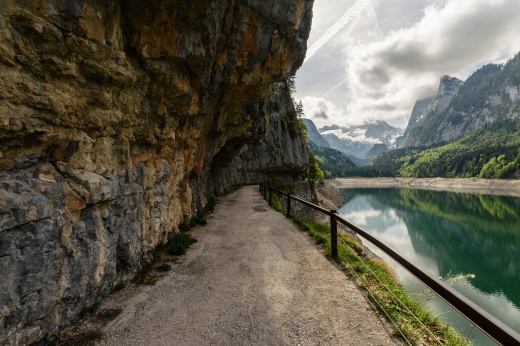 Štěrková cesta se zábradlím pod skalním převisem na břehu Vorderer Gosausee