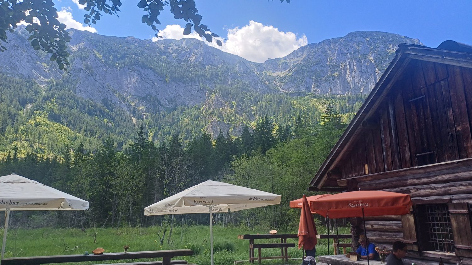 Terasa chaty Taferlklausstub’n se slunečníky a hosty, za ní louka a skály Höllengebirge