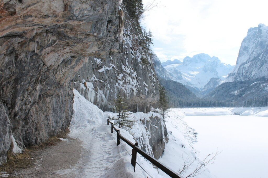 Zasněžená cesta se zábradlím pod skálou nad zamrzlým Vorderer Gosausee, vzadu Dachstein